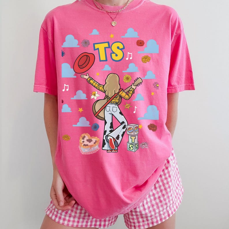 May include: A bright pink t-shirt featuring a graphic of a person with blonde hair playing an acoustic guitar, wearing a yellow plaid shirt and cow-print trousers. The design includes the text TS, a red hat, blue clouds, flowers, and blocks.