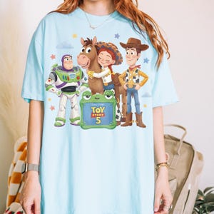 May include: Light blue t-shirt featuring a graphic of characters from Toy Story 5, including Buzz Lightyear, Woody, Jessie, and a horse. The design includes a frog-shaped sign with the movie title and number 5. Stars and clouds are also present.