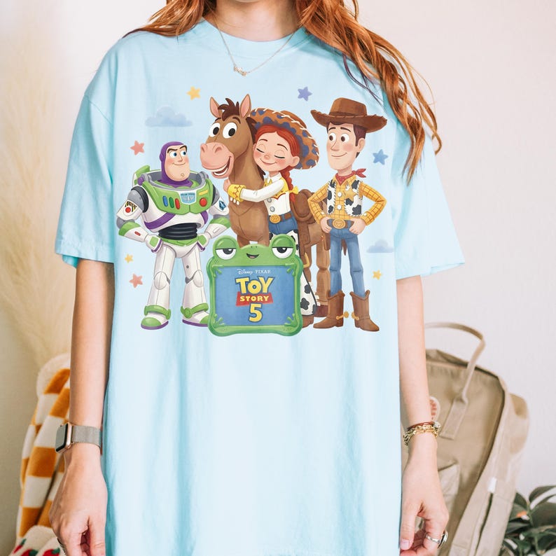 May include: Light blue t-shirt featuring a graphic of characters from Toy Story 5, including Buzz Lightyear, Woody, Jessie, and a horse. The design includes a frog-shaped sign with the movie title and number 5. Stars and clouds are also present.