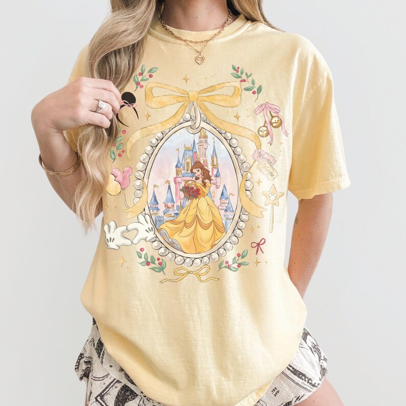 Pu&ograve; includere: T-shirt giallo chiaro con un'illustrazione incorniciata di una principessa in abito giallo che tiene dei fiori davanti a un castello. La cornice &egrave; decorata con fiocchi, perle ed elementi floreali. Sono inclusi dettagli di personaggi dei cartoni animati.