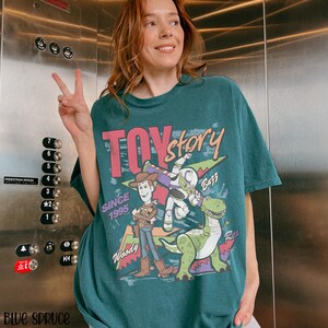 Pode incluir: T-shirt verde-azulado com a inscri&ccedil;&atilde;o "Toy Story" em rosa e branco, com gr&aacute;ficos de Woody, Buzz Lightyear e Rex. A camisa tamb&eacute;m inclui o texto "Since 1995".