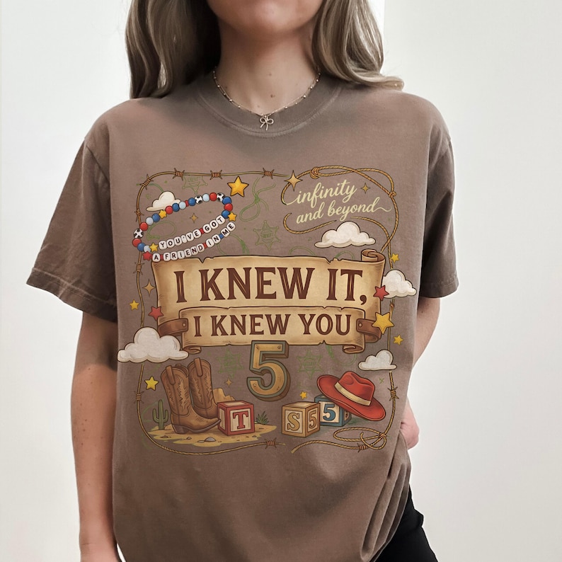May include: A brown short-sleeve t-shirt featuring a graphic print with the text "I KNEW IT, I KNEW YOU", "5", "infinity and beyond", and "YOU'VE GOT A FRIEND IN ME". The design includes cowboy boots, a red hat, alphabet blocks, and a lasso.