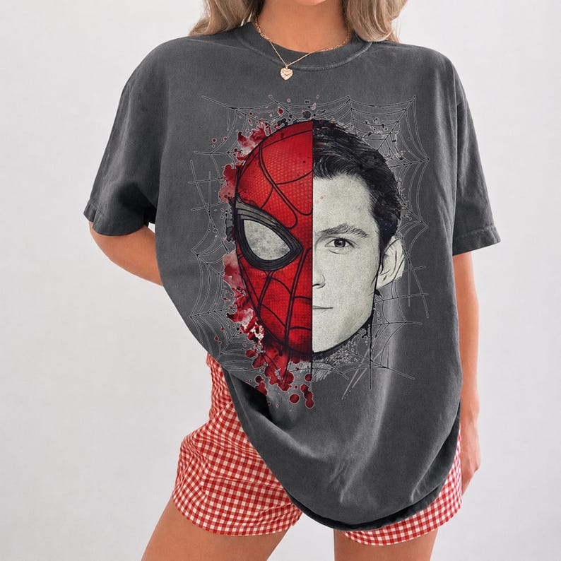 May include: A charcoal gray short-sleeve t-shirt featuring a split-face graphic print. The left side displays a red and black Spider-Man mask, while the right side shows a black and white portrait of a man, all set against a spiderweb background.