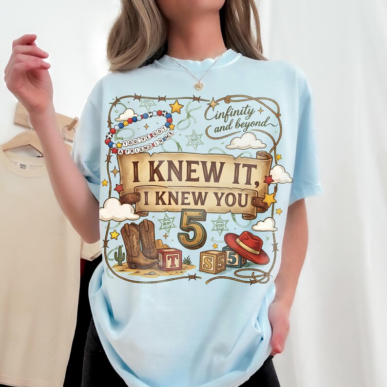 May include: A light blue t-shirt featuring a graphic print with a lasso border, cowboy boots, a red hat, alphabet blocks, and the text "Cinfinity and beyond", "I KNEW IT, I KNEW YOU", "5", and "YOU'VE GOT A FRIEND IN ME" on a beaded bracelet.