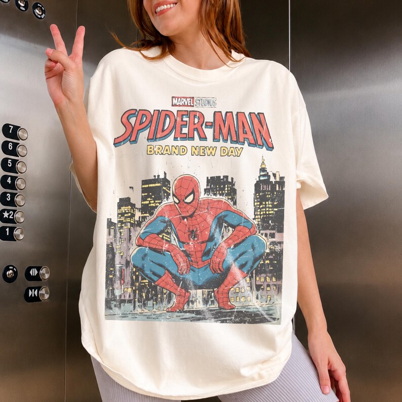 May include: An off-white short-sleeve t-shirt featuring a graphic print of Spider-Man crouching in front of a city skyline. The text reads Marvel Studios Spider-Man Brand New Day. The design has a distressed, weathered appearance.