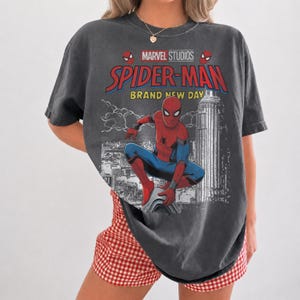 May include: A charcoal gray short-sleeve t-shirt featuring a graphic print of Spider-Man in a crouching pose. The design includes the text Marvel Studios, Spider-Man, and Brand New Day, along with a black and white illustration of a city skyline.
