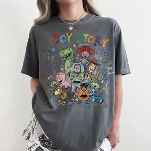 May include: Dark gray t-shirt featuring a colorful graphic of characters from the animated film "Toy Story." The design includes the words "Toy Story" in a playful font, along with drawings of Woody, Buzz Lightyear, and other characters.