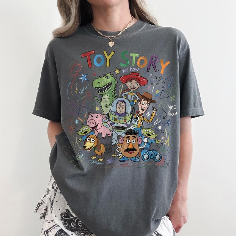 May include: Dark gray t-shirt featuring a colorful graphic of characters from the animated film "Toy Story." The design includes the words "Toy Story" in a playful font, along with drawings of Woody, Buzz Lightyear, and other characters.