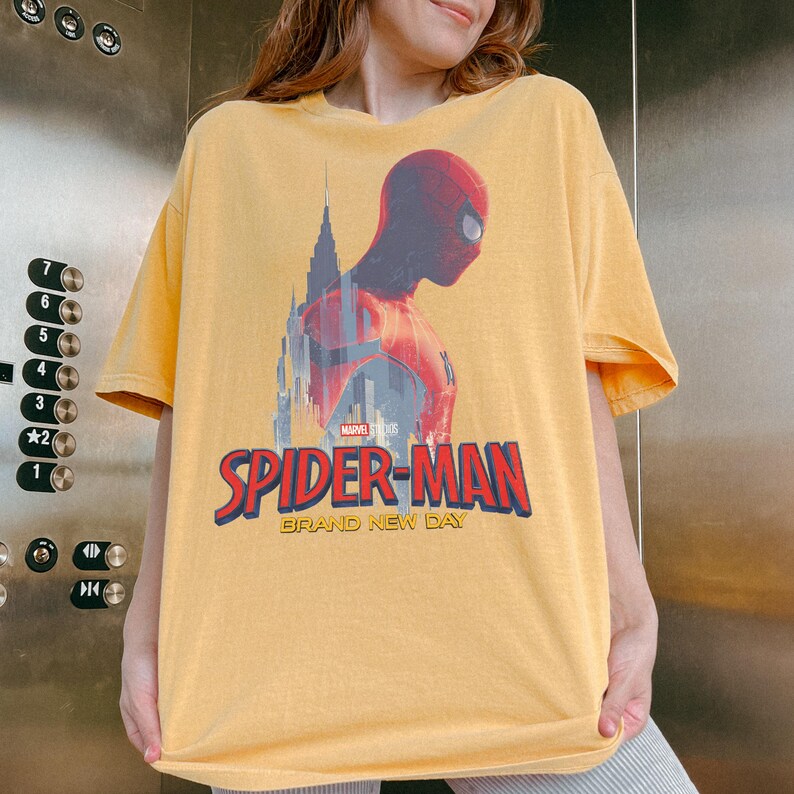 May include: A yellow short-sleeve t-shirt featuring a graphic of Spider-Man in profile against a city skyline. The design includes the Marvel Studios logo and the text "SPIDER-MAN BRAND NEW DAY" in bold red and blue lettering on the front.