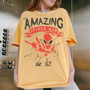 May include: A mustard yellow short-sleeve t-shirt featuring a graphic print of Spider-Man in red and black. The text reads The AMAZING SPIDER-MAN and Est. '62. The design includes a spiderweb pattern and a hand gesture in a distressed style.