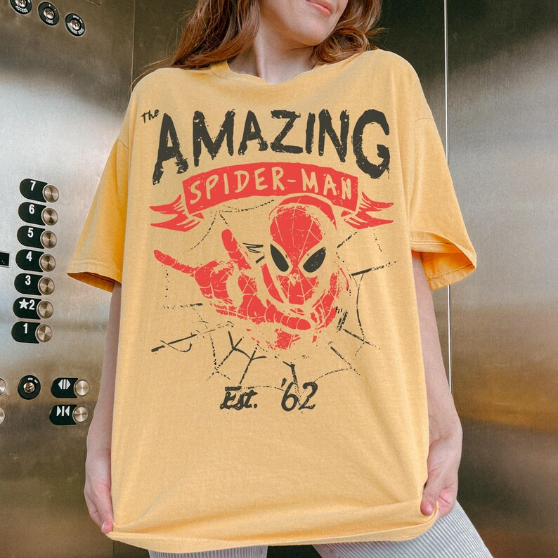 May include: A mustard yellow short-sleeve t-shirt featuring a graphic print of Spider-Man in red and black. The text reads The AMAZING SPIDER-MAN and Est. '62. The design includes a spiderweb pattern and a hand gesture in a distressed style.