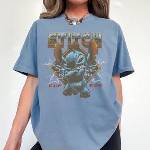 May include: A light blue t-shirt featuring a graphic of the character Stitch from Lilo & Stitch. The word "STITCH" is printed above the character in a retro font. The character is blue with arms crossed, set against a diamond with lightning bolts.