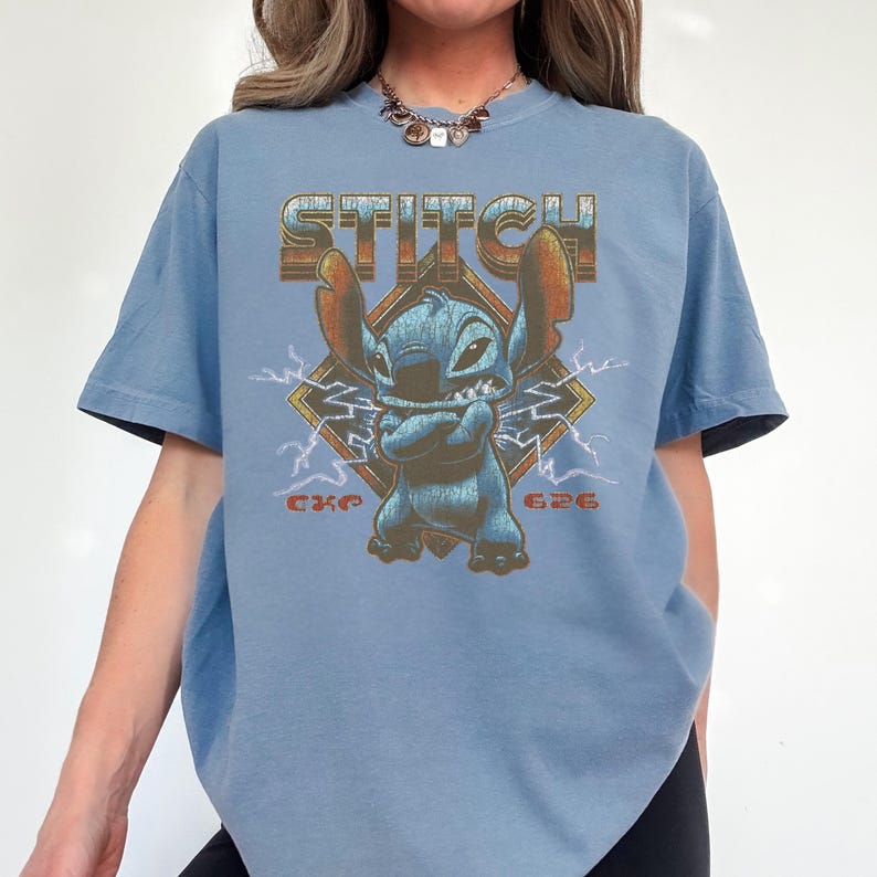 May include: A light blue t-shirt featuring a graphic of the character Stitch from Lilo & Stitch. The word "STITCH" is printed above the character in a retro font. The character is blue with arms crossed, set against a diamond with lightning bolts.