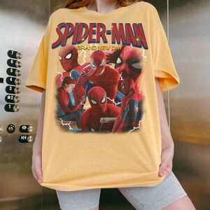 May include: A pale yellow short-sleeve t-shirt featuring a graphic print of Spider-Man in various poses. The text SPIDER-MAN appears in large red and blue letters at the top, with the words BRAND NEW DAY printed in smaller yellow text below.