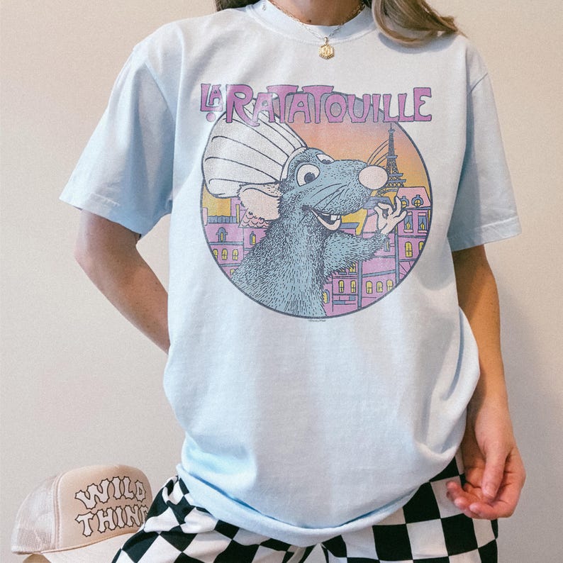 May include: Light blue t-shirt featuring a graphic of Remy from the movie Ratatouille. The design includes the text "Ratatouille" in purple, a cartoon rat, and the Eiffel Tower. The shirt is a crew neck style.