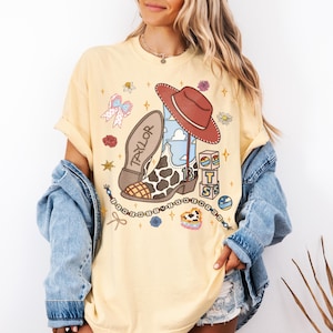 May include: A pale yellow t-shirt featuring a graphic print of a cowboy boot with a cow print pattern, a red hat, and blocks with the letters T, S, and the number 13. The word TAYLOR is printed on the boot sole, surrounded by small icons like bows, flowers, and a beaded string.