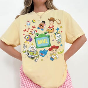 May include: A pale yellow short-sleeve t-shirt featuring a colorful graphic print of various cartoon characters including a cowboy, a space ranger, a cowgirl, a potato figure, and a fork character, with a central green frame containing the text NAME.