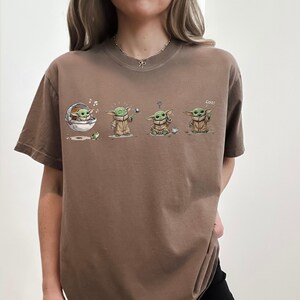 May include: A brown short-sleeve t-shirt featuring a graphic print of a small green creature in four different poses. The illustrations show the character in a floating pod, reaching for a butterfly, sitting with a question mark, and waving with the text Coo!