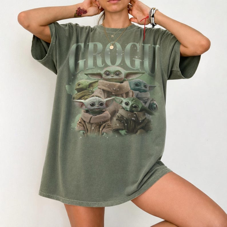 May include: Olive green t-shirt featuring the word "GROGU" in a faded green font. The shirt displays a graphic of Grogu, also known as Baby Yoda, surrounded by other Grogu characters. The shirt has short sleeves and a relaxed fit.