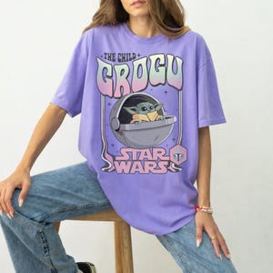 K&ouml;nnte beinhalten: Ein hellviolettes T-Shirt mit einem Grafikdruck der Figur Grogu aus Star Wars in einer schwebenden Kapsel. Der Text lautet THE CHILD GROGU und STAR WARS in einer stilisierten Schrift mit rosa und blauen Farbverl&auml;ufen, erg&auml;nzt durch kleine Sterne.
