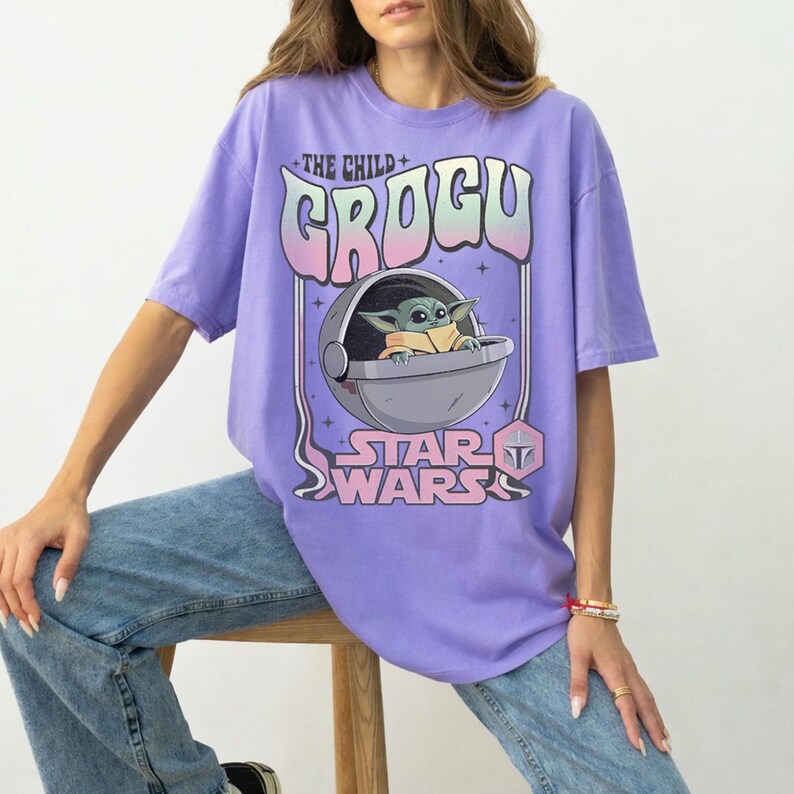 K&ouml;nnte beinhalten: Ein hellviolettes T-Shirt mit einem Grafikdruck der Figur Grogu aus Star Wars in einer schwebenden Kapsel. Der Text lautet THE CHILD GROGU und STAR WARS in einer stilisierten Schrift mit rosa und blauen Farbverl&auml;ufen, erg&auml;nzt durch kleine Sterne.