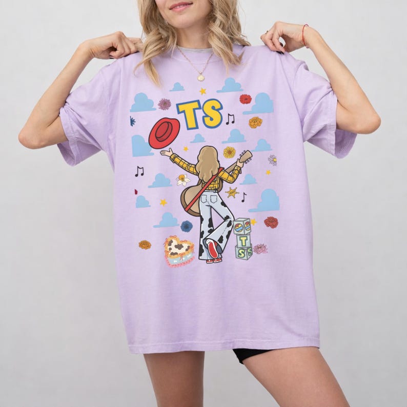 May include: A light purple t-shirt featuring a graphic illustration of a person with blonde hair wearing a yellow plaid shirt and cow print trousers, holding a guitar. The design includes a red hat, blue clouds, flowers, music notes, and blocks with TS.
