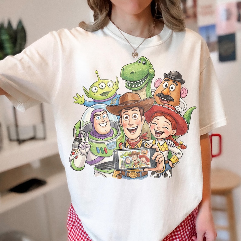 May include: White t-shirt featuring a colorful graphic of Toy Story characters taking a selfie. The design includes Woody, Buzz Lightyear, Rex, Jessie, Mr. Potato Head, and an alien. The shirt has a crew neck and short sleeves.