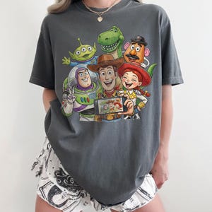 May include: Dark grey t-shirt featuring a colorful graphic of Toy Story characters taking a selfie. The design includes Woody, Buzz Lightyear, Rex, Mr. Potato Head, Jessie, and an alien. The shirt is a comfortable, oversized fit.