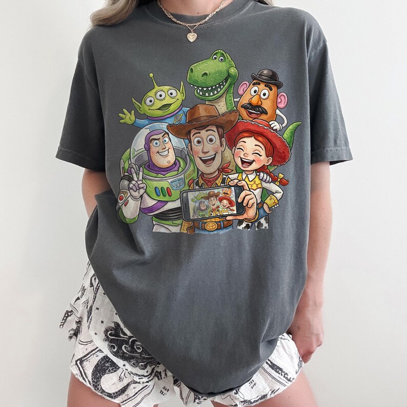 May include: Dark grey t-shirt featuring a colorful graphic of Toy Story characters taking a selfie. The design includes Woody, Buzz Lightyear, Rex, Mr. Potato Head, Jessie, and an alien. The shirt is a comfortable, oversized fit.