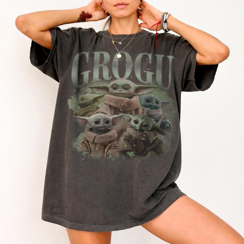 May include: Dark gray t-shirt featuring the word "GROGU" above a graphic of several Grogu characters. The Grogu characters are in various poses and colors, with a soft, faded appearance. The shirt has a vintage, worn look.