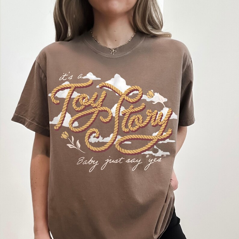 May include: A brown short-sleeve t-shirt featuring a graphic design of white clouds and yellow rope-textured lettering that reads "it's a Toy Story" and "Baby just say yes" in white cursive script, accented with small yellow flower illustrations.