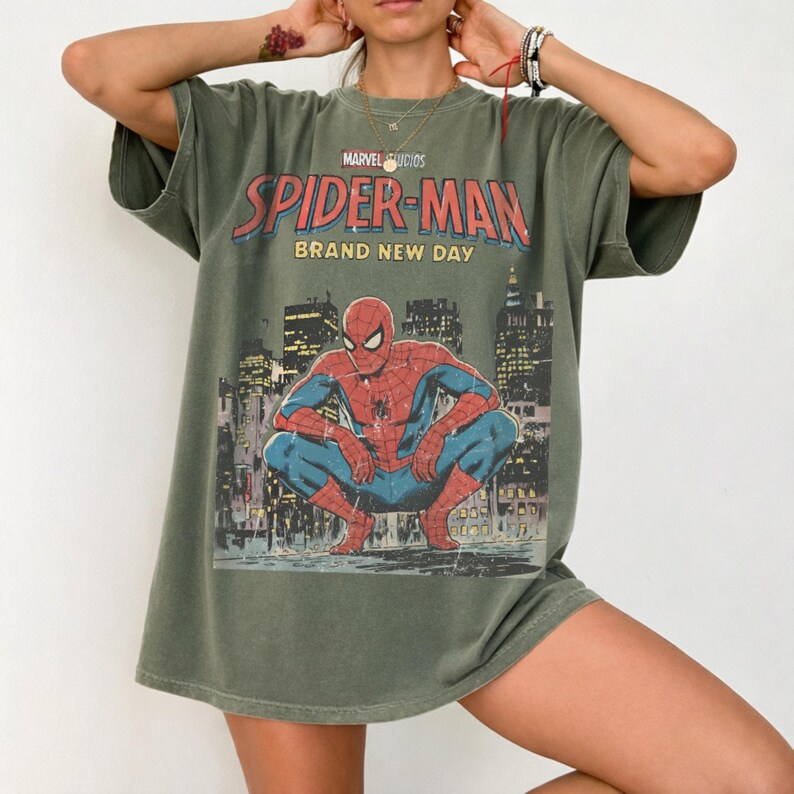 May include: An olive green short-sleeve t-shirt featuring a graphic print of Spider-Man crouching in a city setting. The text reads Marvel Studios Spider-Man Brand New Day. The shirt has a crew neckline and a relaxed, loose-fitting silhouette.