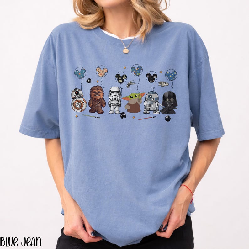 May include: A light blue t-shirt featuring a graphic of Star Wars characters holding balloons. The characters include BB-8, Chewbacca, a Stormtrooper, Grogu, R2-D2, and Darth Vader. The text "BLUE JEAN" is printed at the bottom of the shirt.