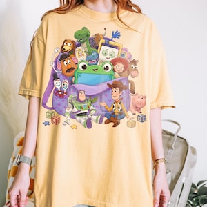 May include: Yellow t-shirt featuring a colorful graphic of characters from the animated film Toy Story. The design includes Woody, Buzz Lightyear, and other beloved characters, with a purple backpack in the center.