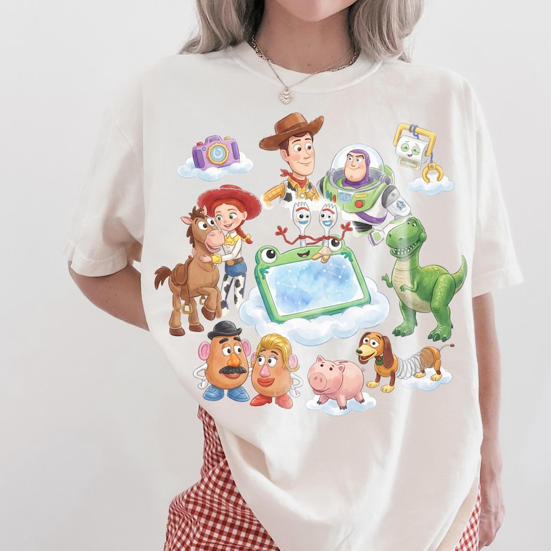 May include: Cream-colored t-shirt featuring a colorful illustration of characters from the Toy Story movie franchise. The design includes Woody, Buzz Lightyear, Jessie, and other beloved characters. The shirt is made of a soft material.