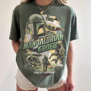May include: Olive green t-shirt featuring a graphic from the Star Wars series, "The Mandalorian and Grogu." The design includes images of the Mandalorian, Grogu, and the text "This is the Way."