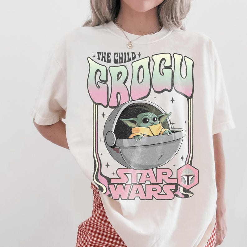 K&ouml;nnte beinhalten: Ein cremefarbenes Kurzarm-T-Shirt mit einem Grafikdruck von Grogu in einer schwebenden Kapsel. Der Text lautet THE CHILD GROGU und STAR WARS, erg&auml;nzt durch ein kleines mandalorianisches Helm-Symbol in pastellgr&uuml;nen und rosafarbenen Akzenten.