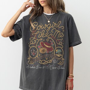 May include: A charcoal gray short-sleeve t-shirt featuring a graphic print with the text Cowgirl Like Me and Takes One to Know One. The design includes a red hat, a horseshoe, a rope border, stars, and small floral accents in muted tones.