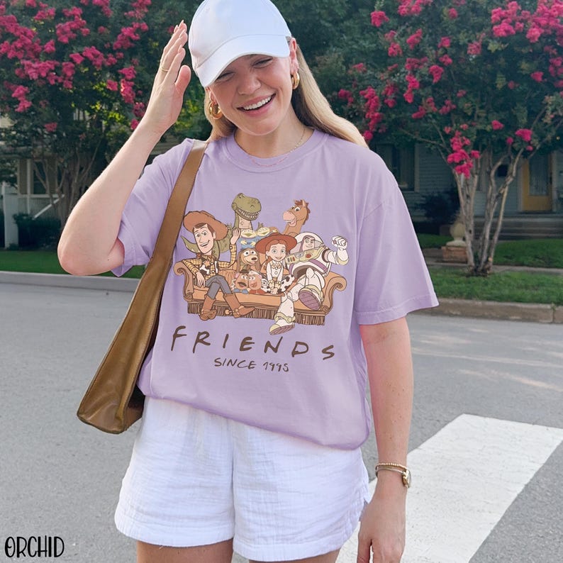May include: A lilac t-shirt with a graphic of "Toy Story" characters on a sofa. The word "FRIENDS" is printed below the graphic, with "SINCE 1995" underneath. The shirt is worn with white shorts. The image is taken outdoors.