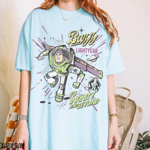 Puede incluir: Camiseta azul claro con un dise&ntilde;o de Buzz Lightyear. El dise&ntilde;o incluye el texto "Buzz Lightyear" y "To Infinity and Beyond". La imagen de dibujos animados es en verde, blanco, morado y negro. Una camiseta divertida e informal.