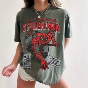 May include: An olive green t-shirt featuring a distressed graphic of Spider-Man in red and black. The text reads "the AMAZING SPIDER-MAN" and includes a small box with "FEATURING PETER PARKER ALIAS SPIDER-MAN" and a box with "ANOTHER SPECTACULAR SMASH-HIT FROM THE HOUSE OF IDEAS!"