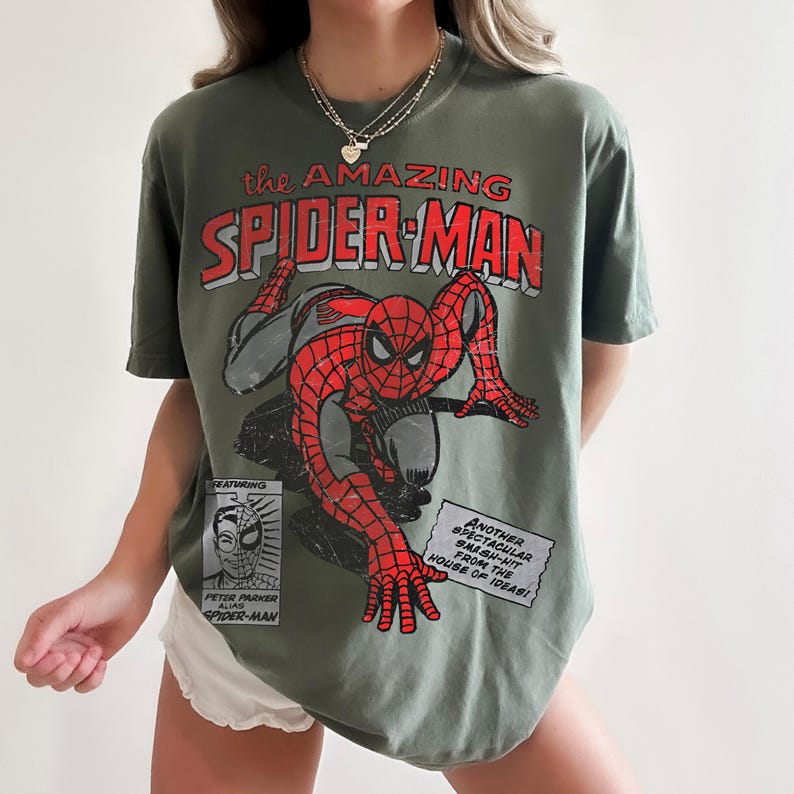 May include: An olive green t-shirt featuring a distressed graphic of Spider-Man in red and black. The text reads "the AMAZING SPIDER-MAN" and includes a small box with "FEATURING PETER PARKER ALIAS SPIDER-MAN" and a box with "ANOTHER SPECTACULAR SMASH-HIT FROM THE HOUSE OF IDEAS!"