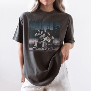以下が含まれることがあります： 暗い灰色のTシャツ。2人の漫画のキャラクター、宇宙レンジャーとカウボーイがベンチに座っているかすれたプリント。画像はヴィンテージルックです。
