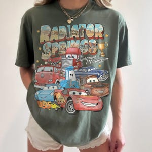Op de afbeelding: Een olijfgroen t-shirt met korte mouwen en een grafische print van Radiator Springs uit de film Cars. Het ontwerp bevat Lightning McQueen, Mater en andere personages, met de tekst Radiator Springs en Lightning McQueen.