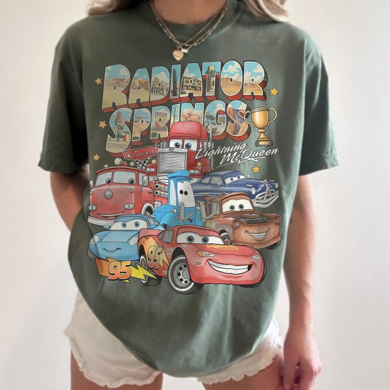 Op de afbeelding: Een olijfgroen t-shirt met korte mouwen en een grafische print van Radiator Springs uit de film Cars. Het ontwerp bevat Lightning McQueen, Mater en andere personages, met de tekst Radiator Springs en Lightning McQueen.