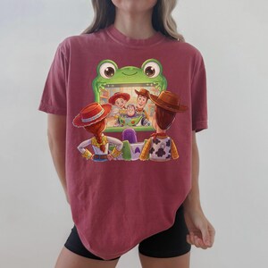 Puede incluir: Una camiseta granate con una pantalla con forma de rana de dibujos animados que muestra personajes de Toy Story. El dise&ntilde;o incluye a Woody, Buzz Lightyear, Jessie y otros personajes. La camiseta es una prenda c&oacute;moda e informal.