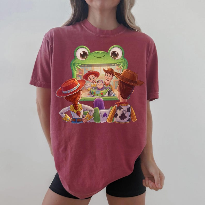 Puede incluir: Una camiseta granate con una pantalla con forma de rana de dibujos animados que muestra personajes de Toy Story. El dise&ntilde;o incluye a Woody, Buzz Lightyear, Jessie y otros personajes. La camiseta es una prenda c&oacute;moda e informal.