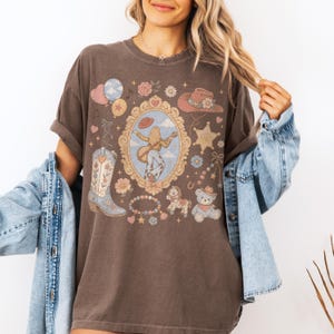 Puede incluir: Una camiseta de manga corta de color marr&oacute;n oscuro con un estampado gr&aacute;fico de un marco dorado ornamentado central que contiene a una persona tocando la guitarra, rodeado por una bota vaquera, un osito de peluche, una estrella y flores.