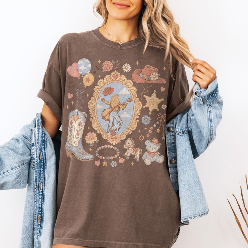 Puede incluir: Una camiseta de manga corta de color marr&oacute;n oscuro con un estampado gr&aacute;fico de un marco dorado ornamentado central que contiene a una persona tocando la guitarra, rodeado por una bota vaquera, un osito de peluche, una estrella y flores.