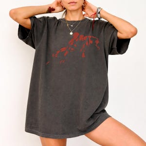 May include: A charcoal grey short-sleeve t-shirt featuring a red graphic print of a figure in a dynamic pose on the chest. The garment has a crew neckline, loose-fitting short sleeves, and a relaxed, oversized silhouette with a faded fabric finish.
